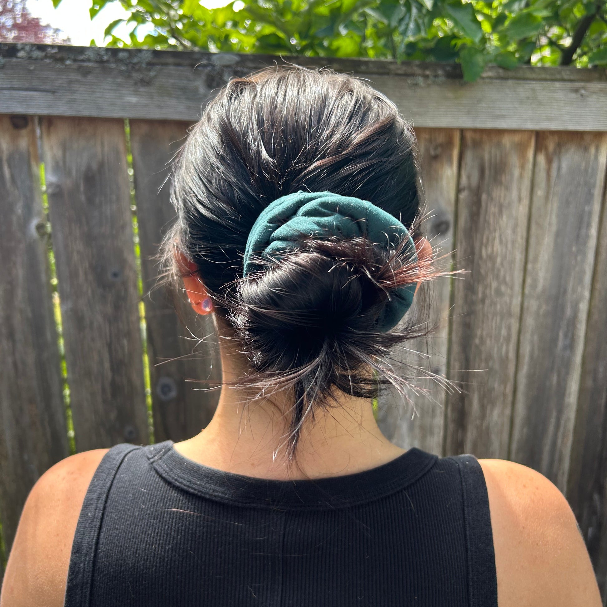Person wearing a black tank top with a green scrunchie, standing outdoors near a wooden fence and greenery.
