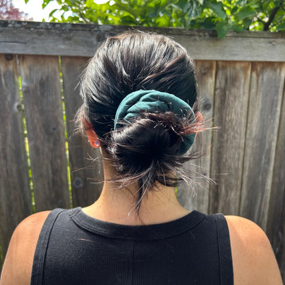 Person wearing a black tank top with a green scrunchie, standing outdoors near a wooden fence and greenery.